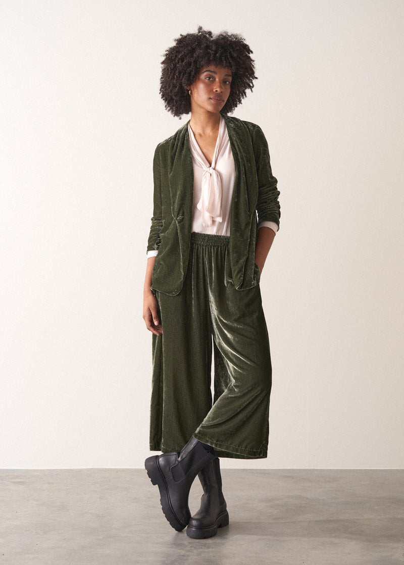 Woman wearing green velvet cropped wide-leg trousers and a pink shirt against a plain background