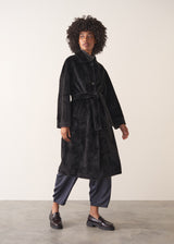 Women wearing an oversized black faux fur coat with blue trousers and loafers, against a beige background