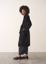 Woman against a beige background, dressed in a long black oversized faux fur coat and jersey bottoms, paired with black loafers.