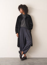 Woman leaning against a beige background, dressed in a long black oversized faux fur coat over a grey sweater and jersey bottoms, paired with black loafers.