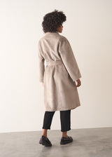 Woman wearing an oversized oatmeal faux fur coat with jeans and loafers, facing a beige background