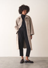 Woman wearing an oversized oatmeal faux fur coat over a black knitted jumper, trousers and loafers, against a beige background