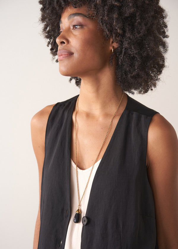 Model wearing a long golden chain with a chunky black oval pendant, layered over a black sleeveless waistcoat, photographed against a simple beige background