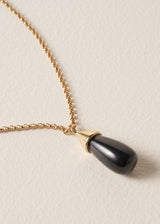 Close up of a long golden chain with a chunky black oval pendant, photographed against a simple beige background