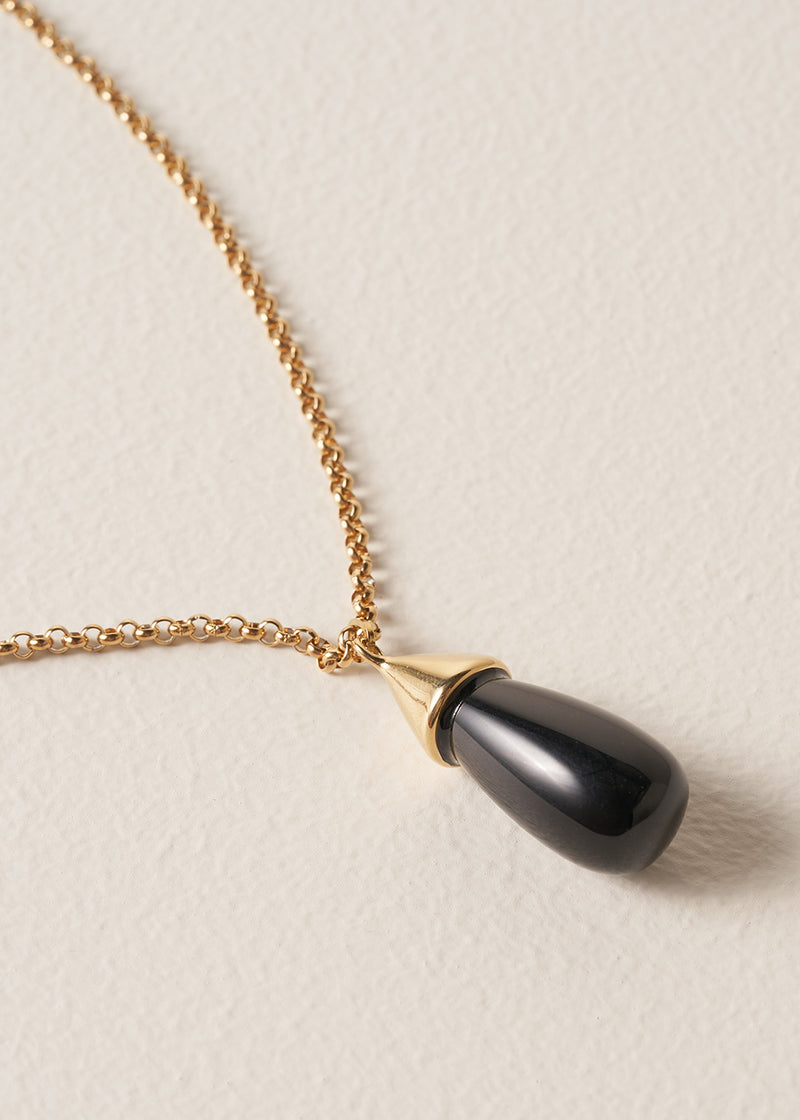 Close up of a long golden chain with a chunky black oval pendant, photographed against a simple beige background