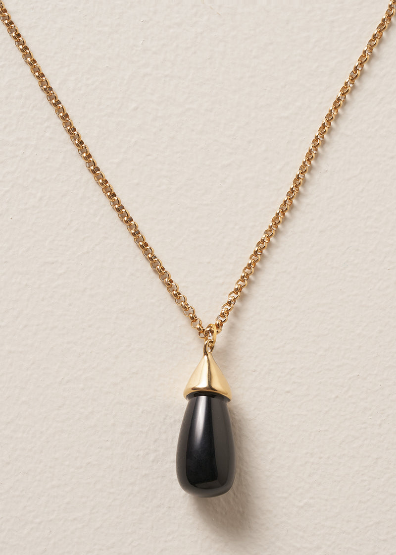 Close up of a long golden chain with a chunky black oval pendant, photographed against a simple beige background