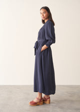 A woman modeling a navy blue maxi dress with long sleeves, a self-tie waist belt, and side pockets, paired with brown platform sandals, set against a neutral studio background.