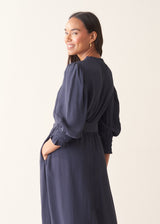 A woman wearing a navy blue midi dress with long balloon sleeves, ruffled cuffs, and side pockets, photographed against a plain light beige studio background.