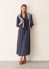 A woman modeling a navy blue maxi dress with a striped cardigan draped over her shoulders, standing against a plain off-white wall.