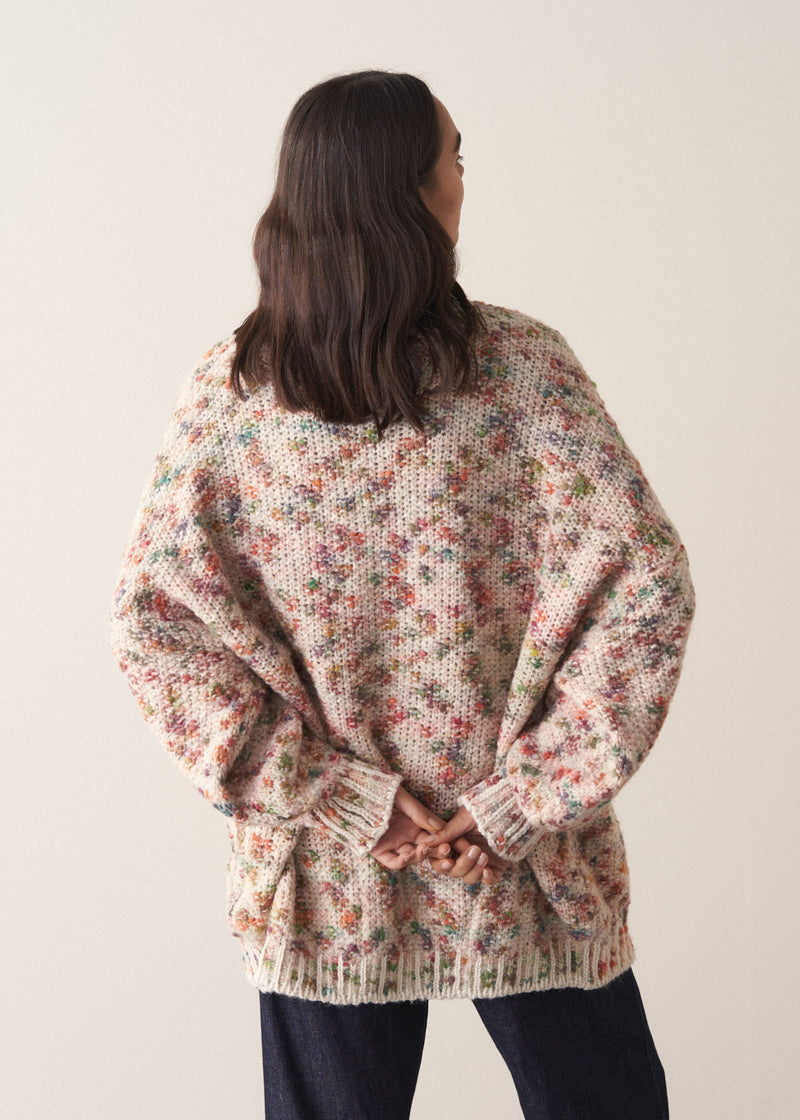 Woman wearing a multicoloured oversized cardigan and jeans  against a plain background
