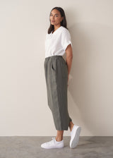 A woman wears a white linen shirt and grey linen cropped barrel-leg trousers with white trainers, against a beige background