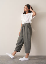 A woman models a white linen shirt and matching linen cropped barrel leg trousers with white trainers, against a beige background