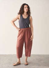 Woman wearing a dark grey tank top and rust-coloured cropped barrel-leg trousers against a plain background