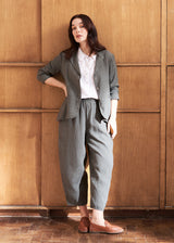 A woman stands against a wooden background, dressed in a khaki linen jacket with matching croppd barrel-leg trousers, a white shirt, and brown loafers