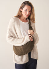 A woman wearing a cream cable-knit cardigan, white t-shirt, and dark blue jeans, carrying an olive green suede crossbody bag against a plain beige background.