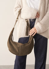 A person wearing a chunky cream knit cardigan, white top, and dark blue jeans carries a slouchy olive-brown suede shoulder bag with a tassel detail.
