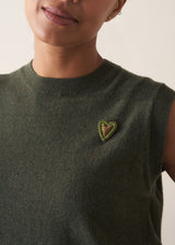Woman wearing a green sleeveless knit with a heart brooch and black trousers against a white background