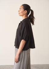 Woman wearing a black linen top and striped, cropped satin trousers against plain background