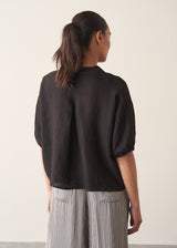 Woman wearing a black linen top and striped, cropped satin trousers against plain background
