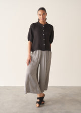 Woman wearing a black linen top and striped, cropped satin trousers against plain background