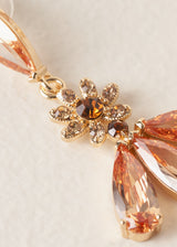 A close-up shot of a drop earring with a floral design and intricate peach-coloured crystals on a beige background