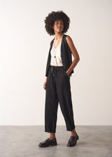 Woman wearing a black waistcoat and black slimfit drawstring trousers against a white wall.