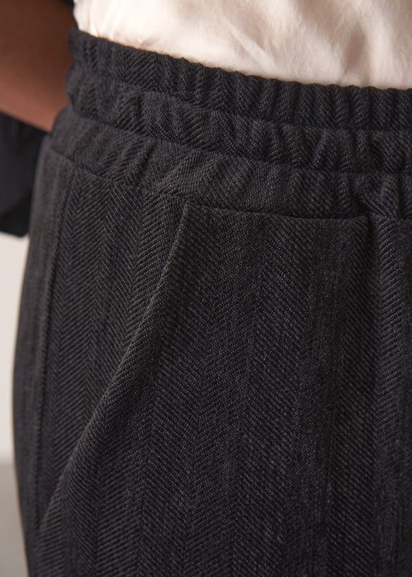 Woman wearing a black waistcoat and black slimfit drawstring trousers against a white wall.