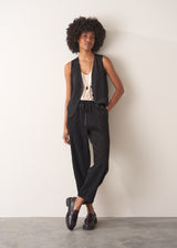 Woman wearing a black waistcoat and black slimfit drawstring trousers against a white wall.
