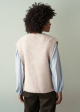 Woman wearing a beige knitted vest over a pale blue shirt and dark green trousers facing a neutral background