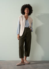 Woman wearing a beige knitted vest over a pale blue shirt and dark green trousers with a neutral background