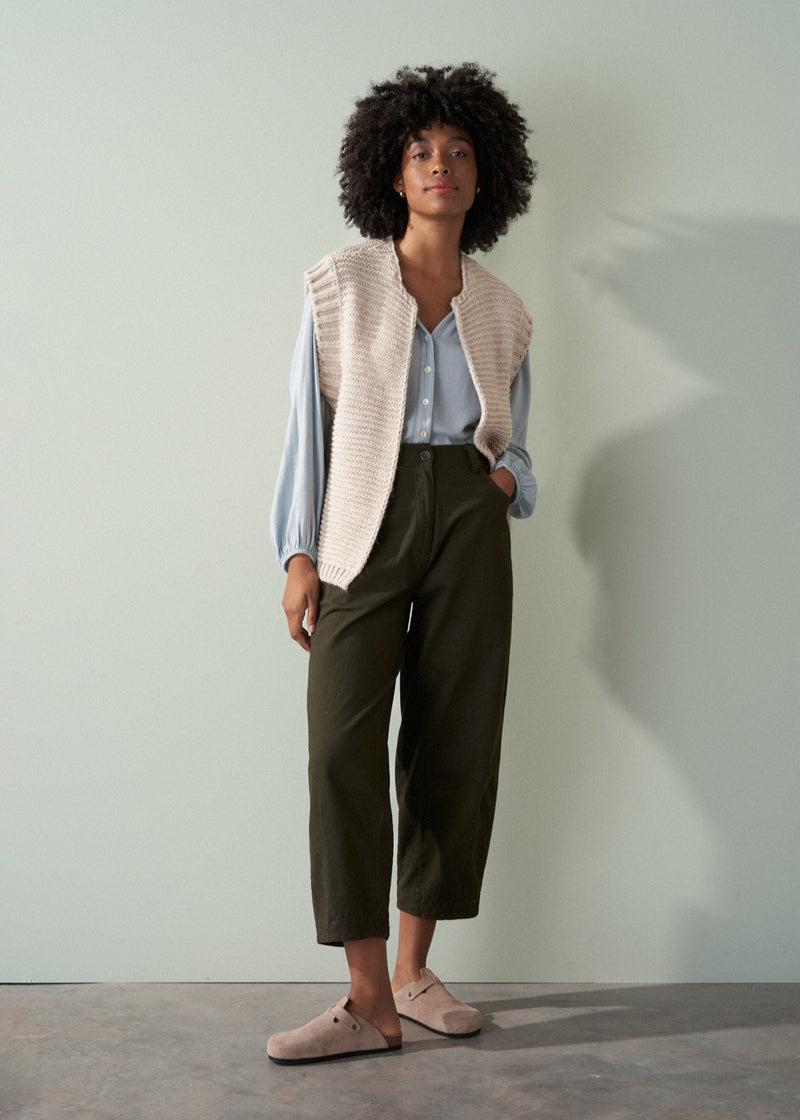 Woman wearing a beige knitted vest over a pale blue shirt and dark green trousers with a neutral background