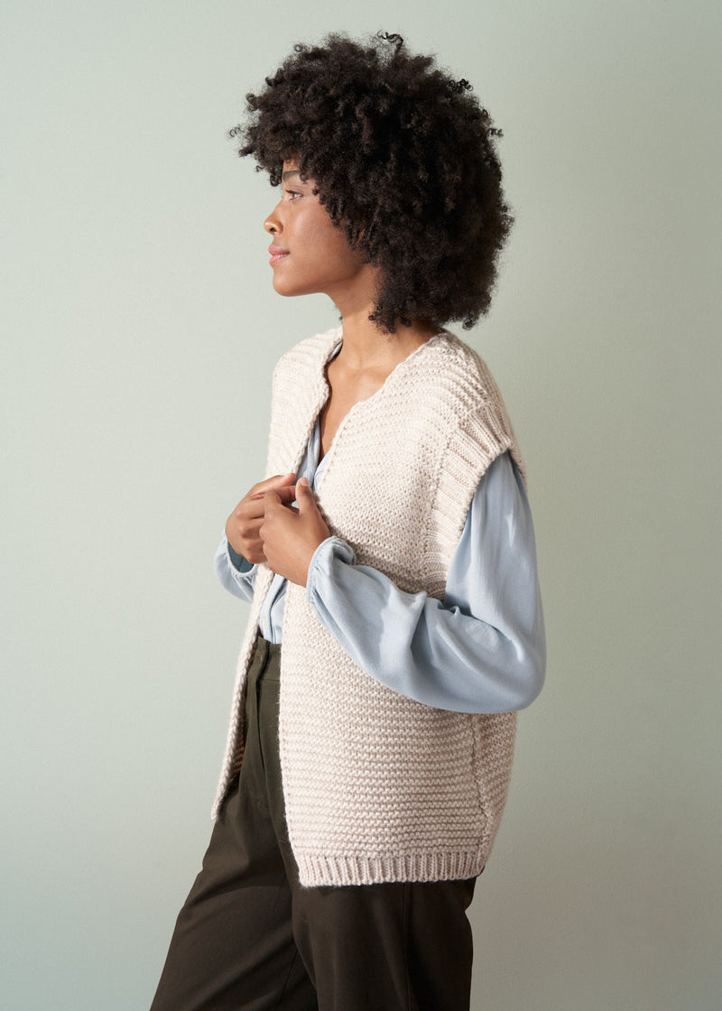 Woman wearing a beige chunky knit waistcoat over a pale blue shirt and dark green trousers facing the side, against neutral backdrop