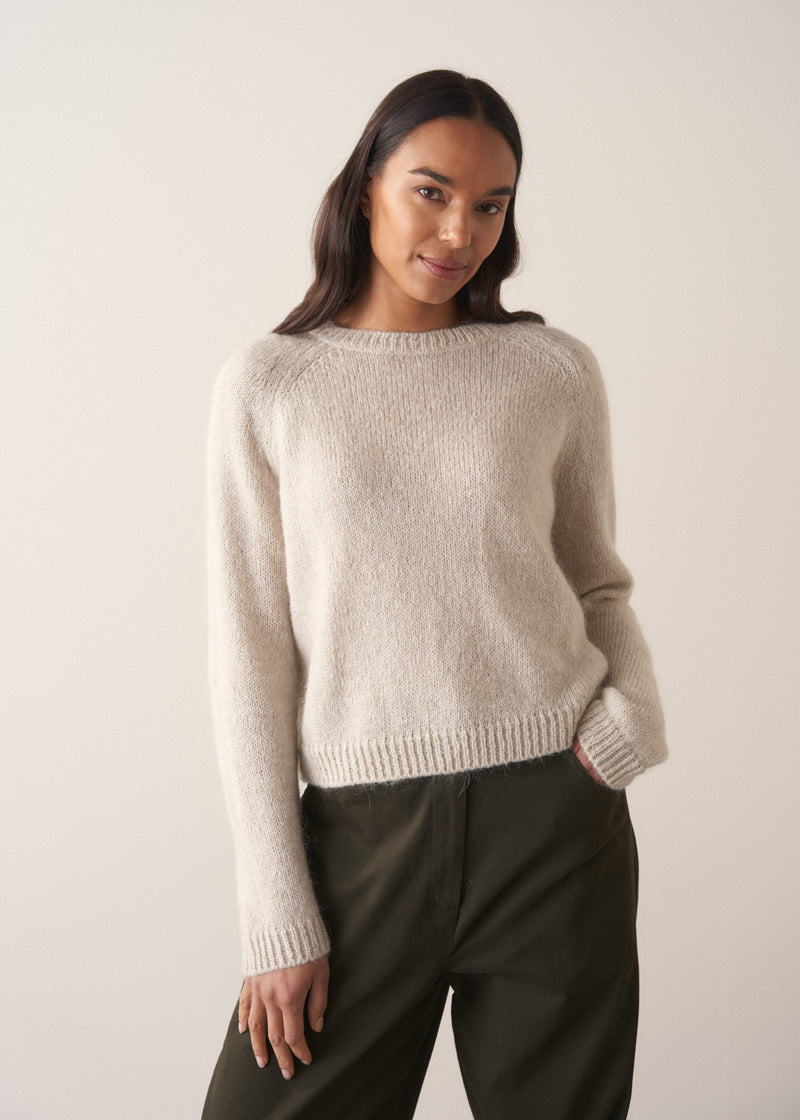 Woman wearing a pale round neck knit sweater with green trousers, against a plain background