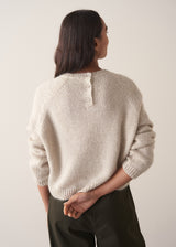 Woman wearing a pale round neck knit sweater with green trousers, facing towards a plain background