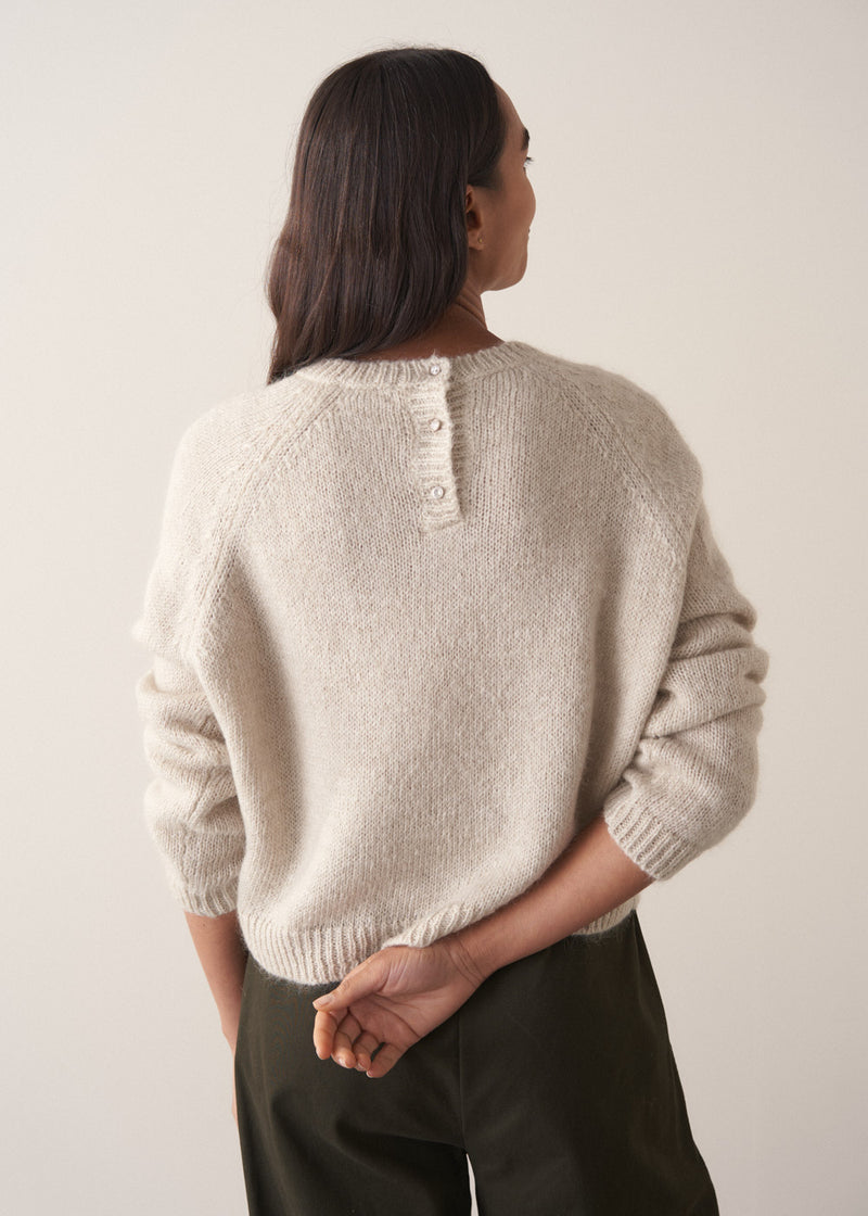 Woman wearing a pale round neck knit sweater with green trousers, facing towards a plain background