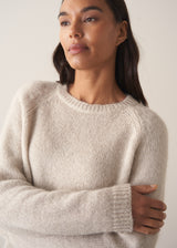 Close up of woman wearing an oatmeal knit round neck sweater against a plain background