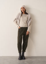 Woman wearing a pale round knit knit sweater with green trousers, against a plain background