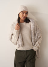 Woman wearing a pale round neck knit sweater with green trousers, against a plain background