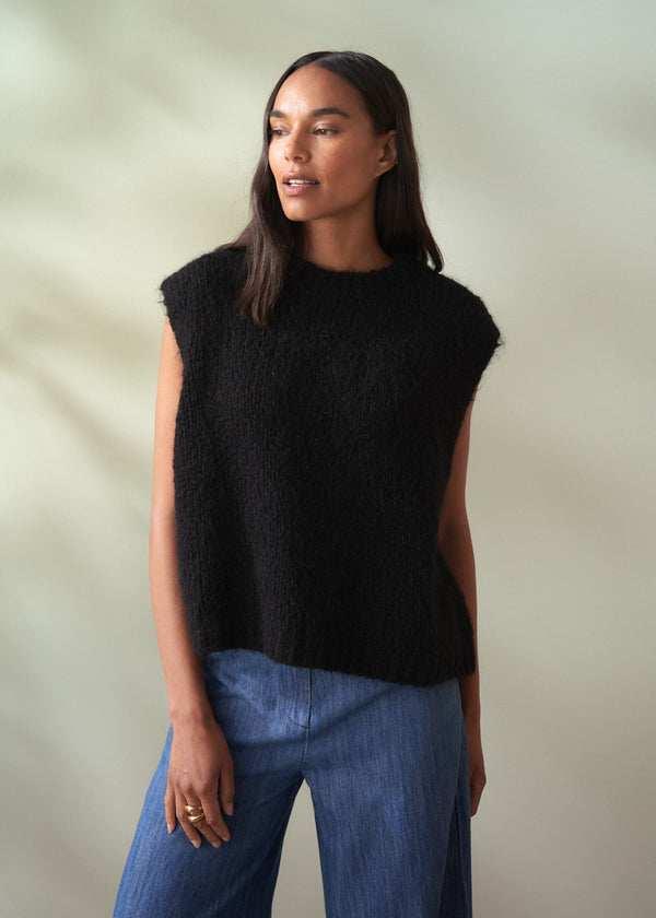 Woman wearing a black sleeveless knit sweater and blue jeans against a neutral background