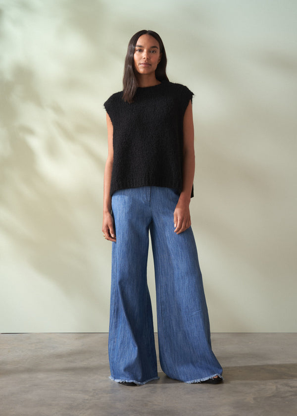 Woman wearing a black sleeveless knit sweater and blue jeans standing against a neutral background