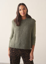 Woman wearing a sage green sleeveless sweater over a long sleeve top and dark brown leopard print trousers against a plain background.