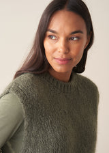 Woman wearing a sage green sleeveless sweater over a long sleeve top and dark brown leopard print trousers against a plain background.
