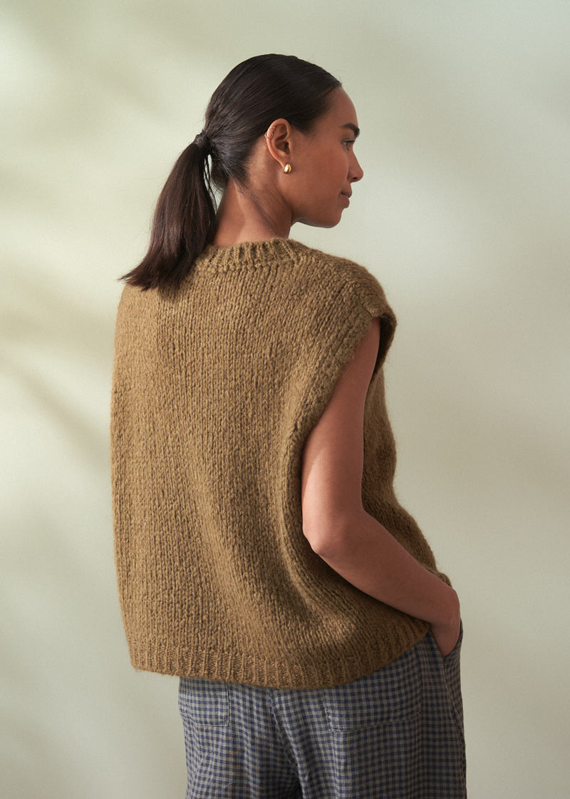 Woman wearing a khaki green knitted vest with check trousers facing a neutral background