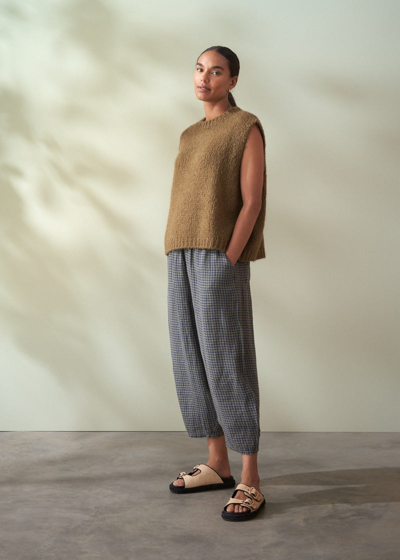 Person wearing a khaki green sleeveless sweater and checkered trousers standing against a neutral background