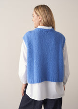 Woman wearing blue knitted sleeveless sweater, a white shirt and dark blue jeans