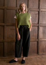 A woman stands in front of a dark wood paneled wall wearing a fuzzy olive green sleeveless top over a white t-shirt, black wide-leg cropped trousers, and black slip-on shoes.