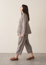 A woman wears a grey and black checked linen jacket paired with matching barrel-leg checked linen trousers and brown loafers. She stands in front of a beige background.