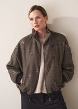 Person wearing a brown textured bomber jacket with sequin detail and black trousers on a plain background