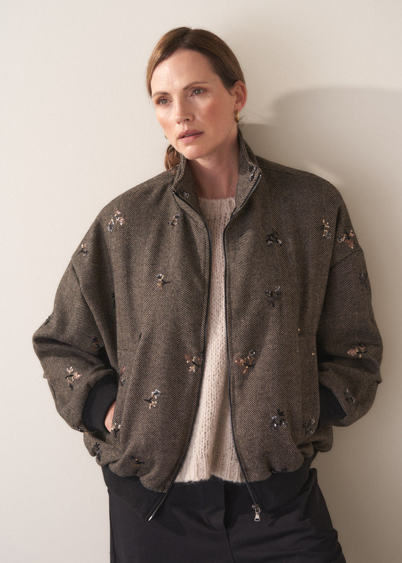 Person wearing a brown textured bomber jacket with sequin detail and black trousers on a plain background