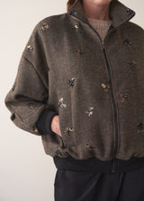 Person wearing a brown textured bomber jacket with sequin detail and black trousers on a plain background
